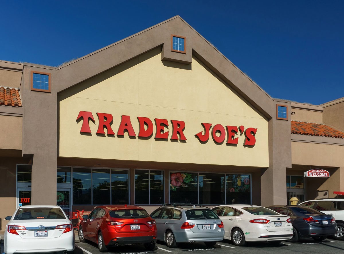 Front view of a Trader Joe's store with a clear blue sky, showing the entrance and a row of parked cars in the lot.