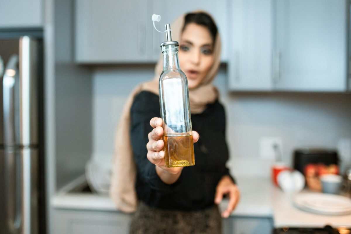 A woman with a headscarf holds a bottle of olive oil in a contemporary kitchen setting.